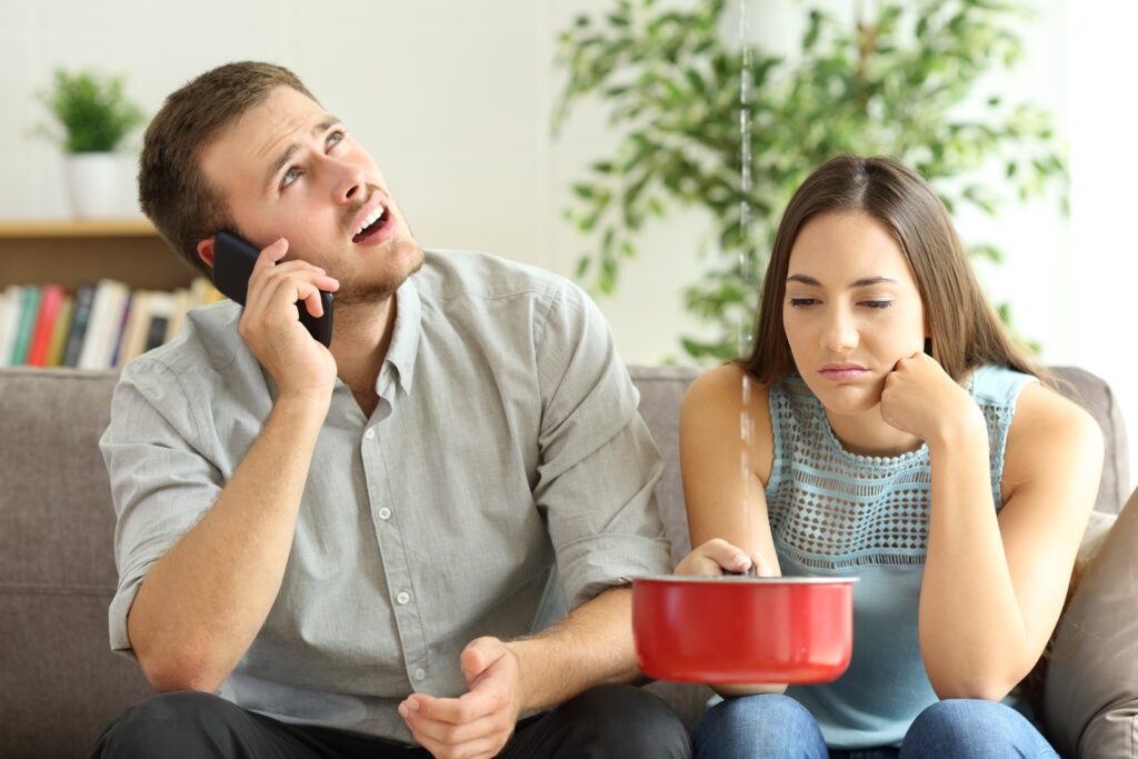 Couple sitting on couch, one catching leaking water in a red pot while the other is looking up at the leak and calling a plumber.