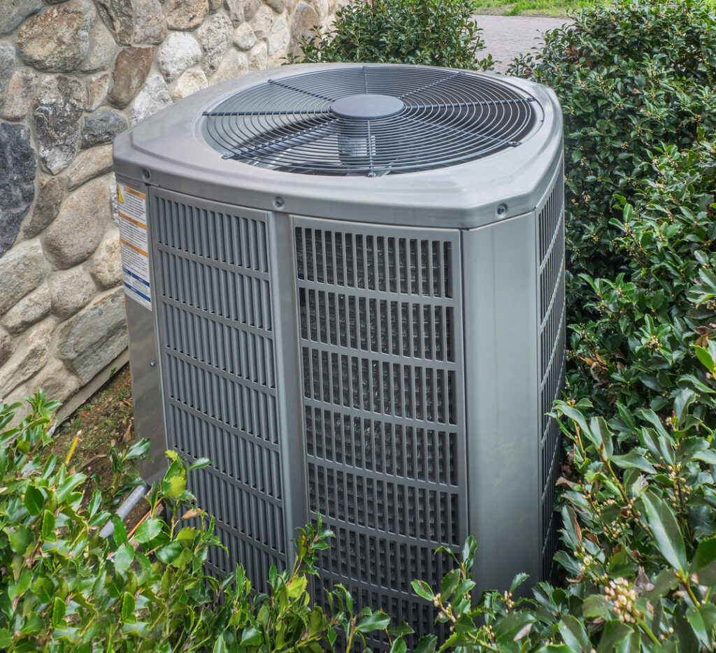 HVAC system outside of a home, next to a stone foundation, with small shrubs surrounding.